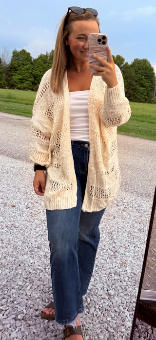 Woman taking a mirror selfie wearing a cream cardigan, white top, and blue jeans outdoors.