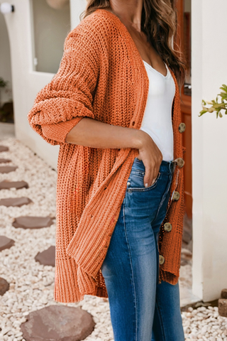 Person wearing an orange knitted cardigan over a white top and blue jeans, standing on a stone path.